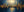 A horizontal hand-painted abstract oil painting showing a shimmering golden city skyline with 3D texture against a deep midnight blue background, reflecting in water.
