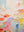 A vertical hand-painted abstract oil painting with thick, sculptural impasto textures in pastel colors like lavender, yellow, orange, and mint green on a cream background.