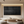 Large horizontal handmade black oil painting with heavy vertical textured ridges, displayed in a warm modern living room above a wooden coffee table with floor cushions.