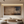 Large horizontal handmade abstract oil painting in deep brown and charcoal with weathered textures, hanging in a modern white dining room above a dark wood table with grey chairs.