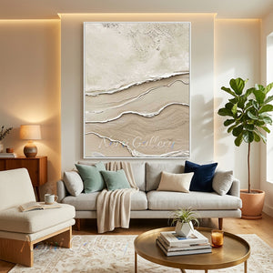 Large handmade 3D abstract canvas art featuring heavy plaster textures of sand ridges and ocean wave reliefs in neutral beige and cream tones, framed in white and displayed on a modern warm-lit living room wall above a grey fabric sofa.