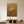Large handmade 3D abstract canvas painting covered entirely in shiny metallic gold leaf, featuring dramatic flowing fluid plaster textures and deep ridges, displayed on a dark grey concrete minimalist living room wall above a modern grey sofa.