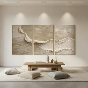 Three-panel hand-painted abstract oil paintings featuring heavy impasto textures that resemble ocean waves and sandy beaches in a minimalist cream and beige color palette, styled in a modern sunlit living room.