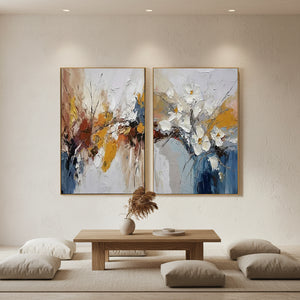 A pair of hand-painted abstract floral oil paintings featuring heavy 3D impasto textures in white, yellow, and navy blue, styled on a neutral wall in a modern sunlit living room.