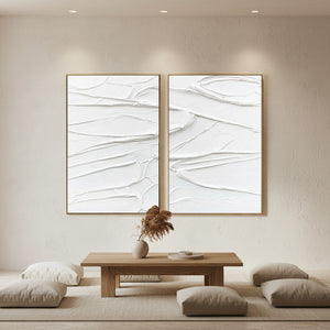 A pair of hand-painted 3D white textured plaster art canvases with bold sculptural ridges, styled in a minimalist sunlit living room above a low wooden table and neutral floor cushions.