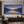 A 100% hand-painted impressionist landscape oil painting featuring deep purple and blue mountains flanking a calm lake that reflects a warm golden sunset sky.