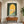 Hand-painted minimalist oil painting showing a woman in a terracotta dress sitting on green steps under a dark olive green archway, looking out towards a bright golden and grey sandy landscape.