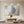 A pair of large hand-painted abstract paintings with grey, beige, and gold leaf textures, displayed in a modern sunlit minimalist living room above a wooden coffee table.