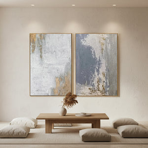 A pair of large hand-painted abstract paintings with grey, beige, and gold leaf textures, displayed in a modern sunlit minimalist living room above a wooden coffee table.