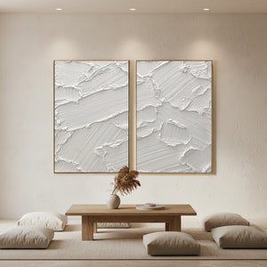 Pair of hand-painted white abstract paintings with heavy diagonal 3D impasto brushstrokes, shown in a modern minimalist living room featuring a wooden table and low seating.