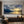 A detailed close-up of a hand-painted impressionistic oil painting on canvas, showing a dramatic sunset with gold and yellow light breaking through deep blue-grey clouds over a tranquil mountain lake. The expressive brushwork and texture of the paint are clearly visible.