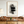 Hand-painted abstract oil painting with two heavy textured black rectangular blocks overlaid with thin intersecting lines, framed in black and styled in a bright, high-ceiling modern home interior next to a minimalist concrete staircase.
