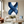 Large handmade abstract oil painting showing a minimalist X-shape intersection of two large textured navy blue brushstrokes on a white canvas, enclosed in a black frame and mounted on a beige home office wall above a sleek white desk with a desk lamp.