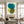 Large handmade abstract painting featuring layered, translucent organic circles in emerald teal, olive green, and mustard yellow on a cream canvas background, framed in black and hung on a minimalist white hallway wall above a dark wood console table next to a staircase.
