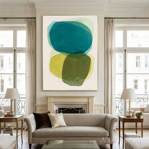 Large handmade abstract painting featuring layered, translucent organic circles in emerald teal, olive green, and mustard yellow on a cream canvas background, framed in black and hung on a minimalist white hallway wall above a dark wood console table next to a staircase.