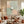 Large handmade abstract canvas painting with layered color blocks of sage green, olive, and warm cream overlapped with fine black and white curved lines, framed in black and mounted on a minimalist wall in a modern living room interior.
